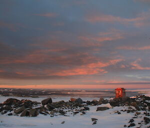 Small cabin on rocky outcrop over looking icebergs in the sea and cloudy sky lit up by sunset