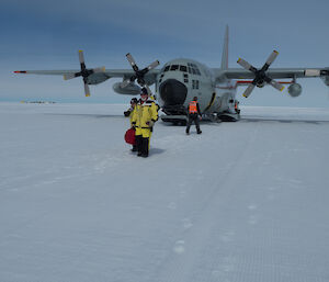 Ice runway, large grey hercules aircraft fills picture, two people in yellow and black antarctic pants and jacket walk from the plane towards the photographer