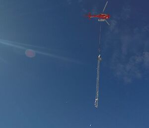 Blue sky fills picture, small red helicopter slinging a large metal mast below
