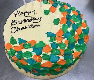 Cream cake with blue orange and green iced smears and brown writting Happy Birthday Charlton