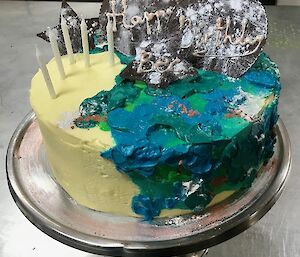 Cream cake with blue and green icing on side and half of the top, four white candles and tempered dark chocolate disc with Happy Birthday Bec written on it