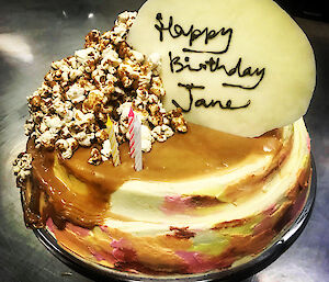 Pink and cream two layered cheese cake with caramel sauce and popcorn spilling off one side. Tempered chocolate disc sitting in cake with Happy Birthday Jane written on it