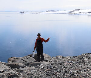 Man standing on rocky foreground and waving, In the background is a large open water bay with snow and rock covered hills beyond