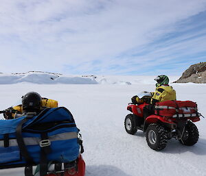 Foreground quad bike up close, middle picture a man with helmet on sitting on red quad bike. On flat ice covered ground. In the distance rocky and snow covered hills.