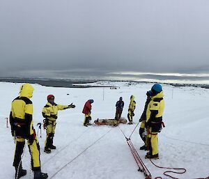 Stretcher connected to rope system with expos on either side listening to instructor. Set up on snowy slope with sea and dark rain clouds in background