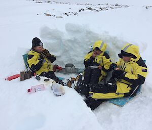 Three expeditioners sitting in a dug out snow hollow eating from mugs with middle expo on the radio reading from a notebook