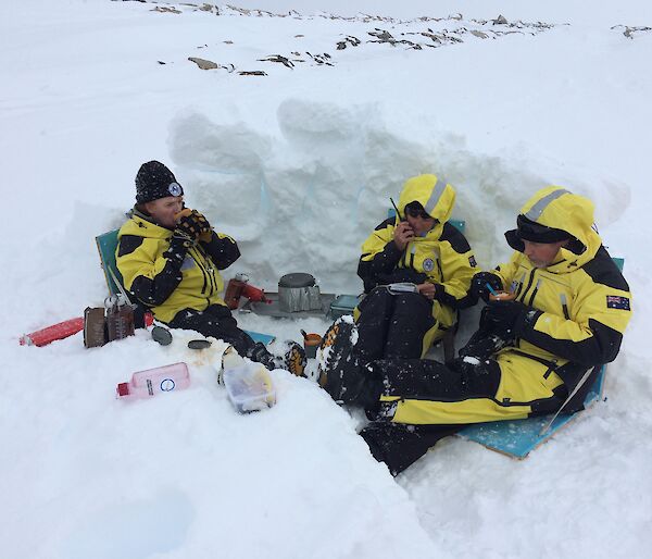 Three expeditioners sitting in a dug out snow hollow eating from mugs with middle expo on the radio reading from a notebook