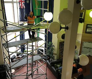 Large double story internal space with pillar and globe lighting at front right, centre and left is a stack of scaffolding with man on top level standing with thumb raised