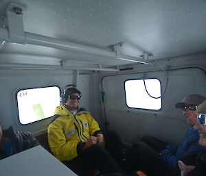 Group of four people sitting in the back of a hagglunds vehicle, white painted walls, two windows with bright light shining in