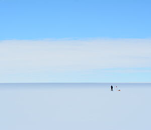 In the middle distance a tiny person standing beside a cane with red flag on top and tools. Foreground to horizen is white ice expanse, above is blue sky with white cloud covering bottom half