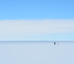 In the middle distance a tiny person standing beside a cane with red flag on top and tools. Foreground to horizon is white ice expanse, above is blue sky with white cloud covering bottom half
