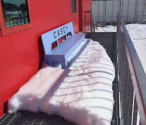 Metal grid deck in front of red building, Blue bench with CASEY cut into the back leaning against building with feet in large snow drift