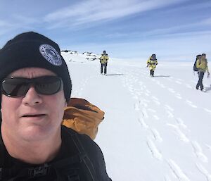Selfie of man in dark sunglasses and black beanie with pack on back in foreground on left of picture. In background, snow covered ground stretching to horizen with blue sky above with sweeping cirostratus clouds. A line of three expeditioners in distrance walking towards the camera