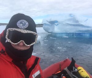 A selfie of a man in red mustang suit, black beanie, neck warmer and goggles, in front left of picture. Bottom right is the bow of the RHIB he is sitting in. In the distance is an iceberg floating in grey-blue water