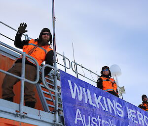 Varandah outside red containerised building, with three men in high vis vest with one closest to the camera waving and smiling. In front a blue sign reading Wilkins Aerodrome
