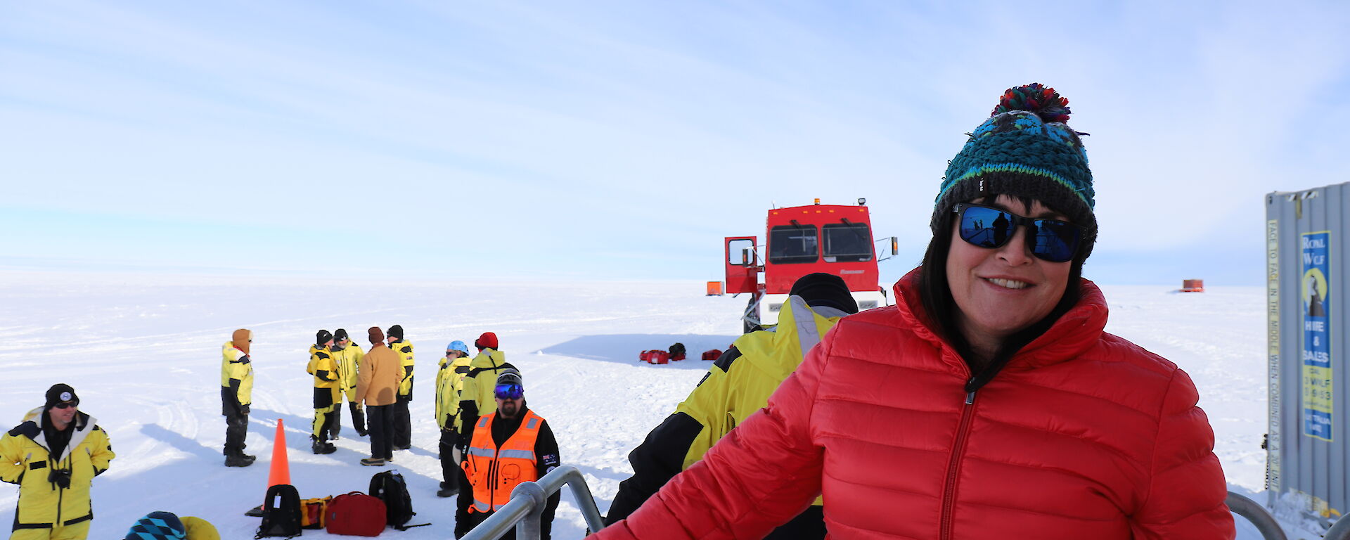 Lady in red jacket and coloured beanie on varandah with group of people and red bus (front on) behind