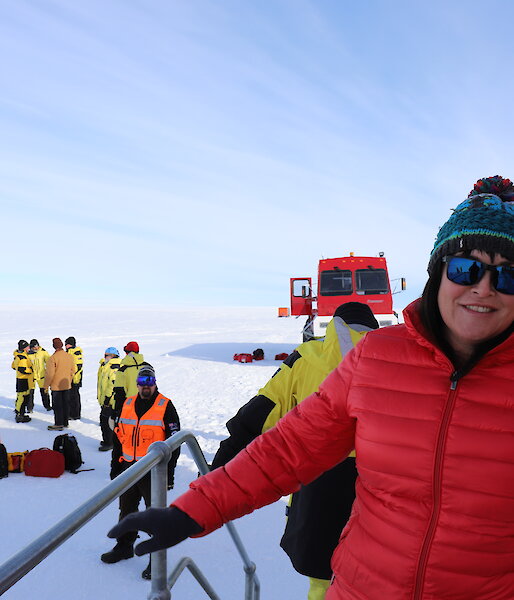 Lady in red jacket and coloured beanie on varandah with group of people and red bus (front on) behind
