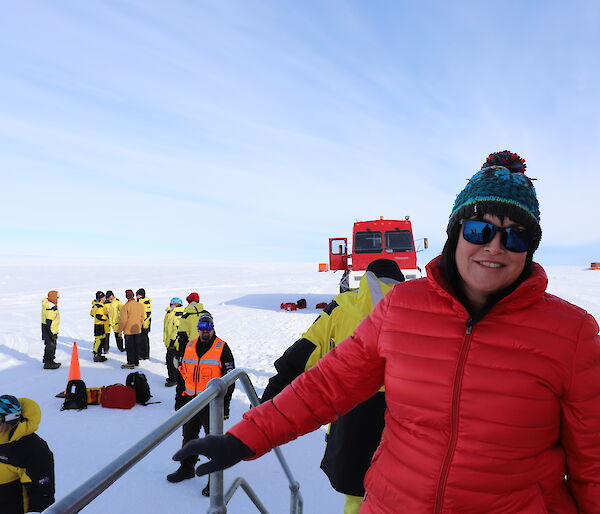 Lady in red jacket and coloured beanie on varandah with group of people and red bus (front on) behind