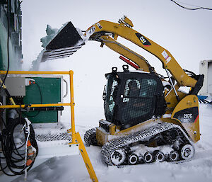 Man in bob cat shovelling snow into a large container attached to side of green building