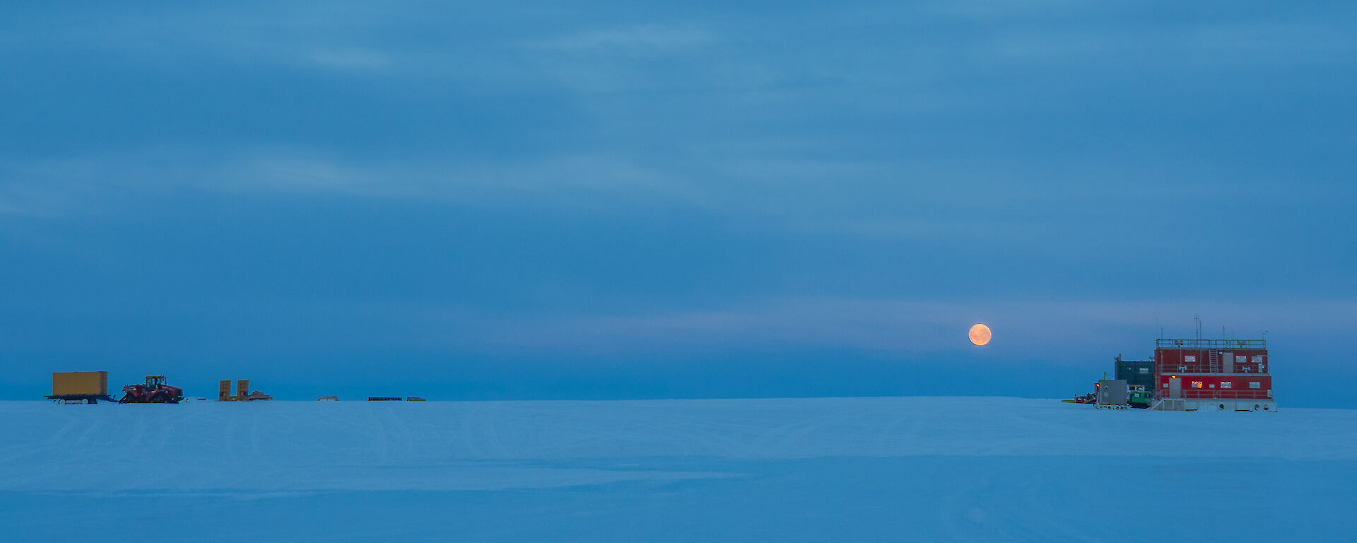 Snow covered ground to horizen and cloud covered sky all a similar hue of purple blue at twighlight, in the distance vehicles are parked and a collection of buildings, and in far distance the full moon just in a dark orange just above the horizen.