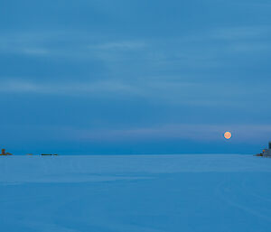 Snow covered ground to horizen and cloud covered sky all a similar hue of purple blue at twighlight, in the distance vehicles are parked and a collection of buildings, and in far distance the full moon just in a dark orange just above the horizen.