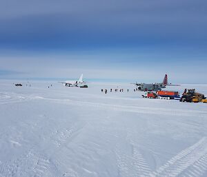 Ice runway with blue sky above, on apron a grey hurcules aircraft and a white A319 aircraft with personnel transfering between