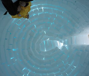 The inside of the igloo looking directly up, the ice blocks have a blue tinge from the outside light, one side is an entry way and on the otherside a person in cold weather gear