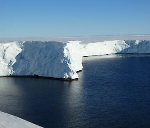 The ice cliffs of a glacier jutting out into a bay of dark blue water; bright blue sky above