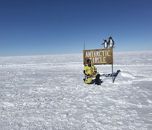 Man kneeling in front of antarctic circle sign, ice covered ground to horizen and bright blue sky above