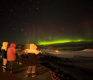 A group of people in cold weather gear photographed from behind, on the distant horizen is a bright green aurora and stary night sky above