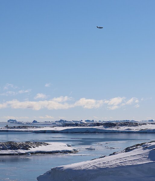 C-17 aircraft flies over Newcomb Bay, blue sky and seas with snow covered rocky shoreline eitherside of bay