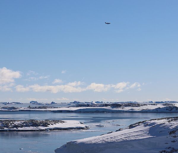 C-17 aircraft flies over Newcomb Bay, blue sky and seas with snow covered rocky shoreline eitherside of bay