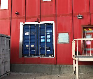Side of a red painted building and in the centre a hole filled with the end of a blue shipping container