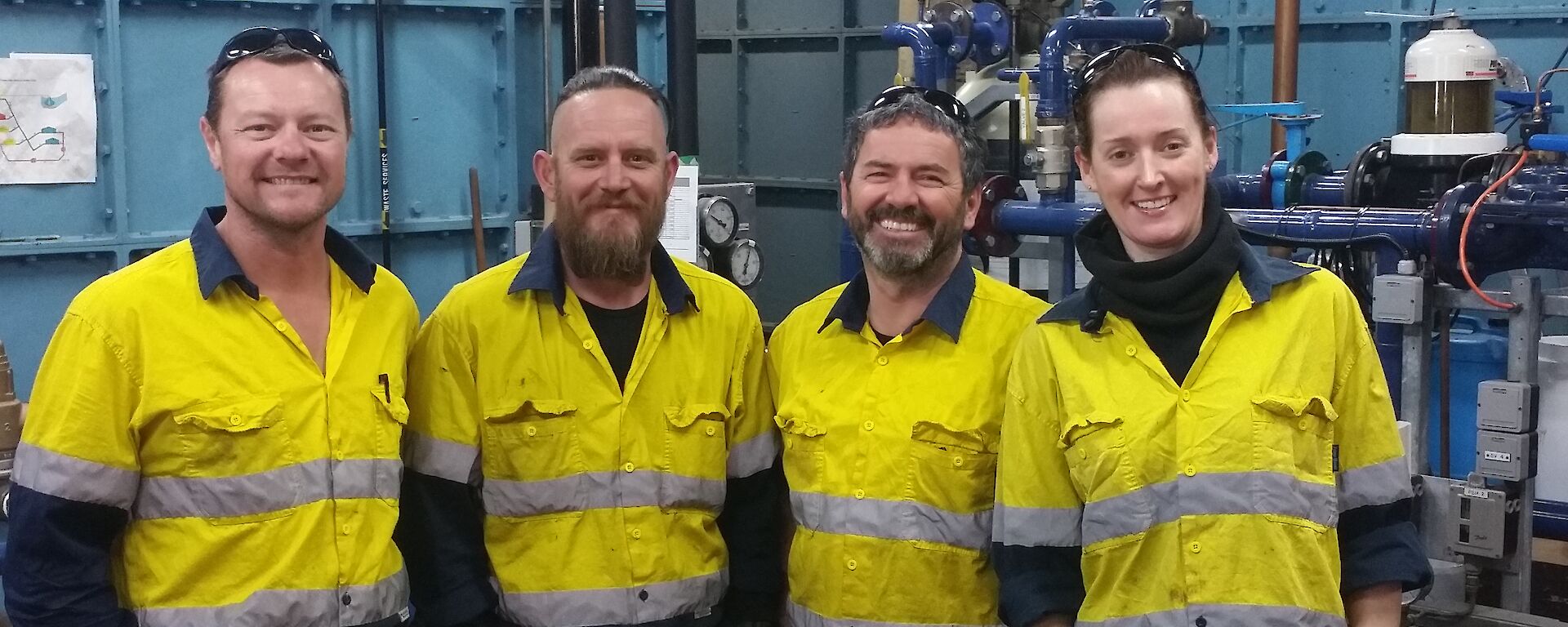 Three men and one woman in high vis smiling to camera with internals of tank house behind