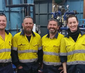 Three men and one woman in high vis smiling to camera with internals of tank house behind