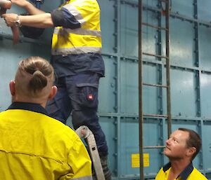 Man up ladder working on piping on side of large tank. Two men standing below watching on.