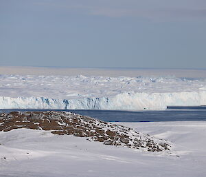 Foreground; snow covered ground with rocky hill. Middle picture; bay with glacier jutting out. Above; blue sky with bands of cloud.