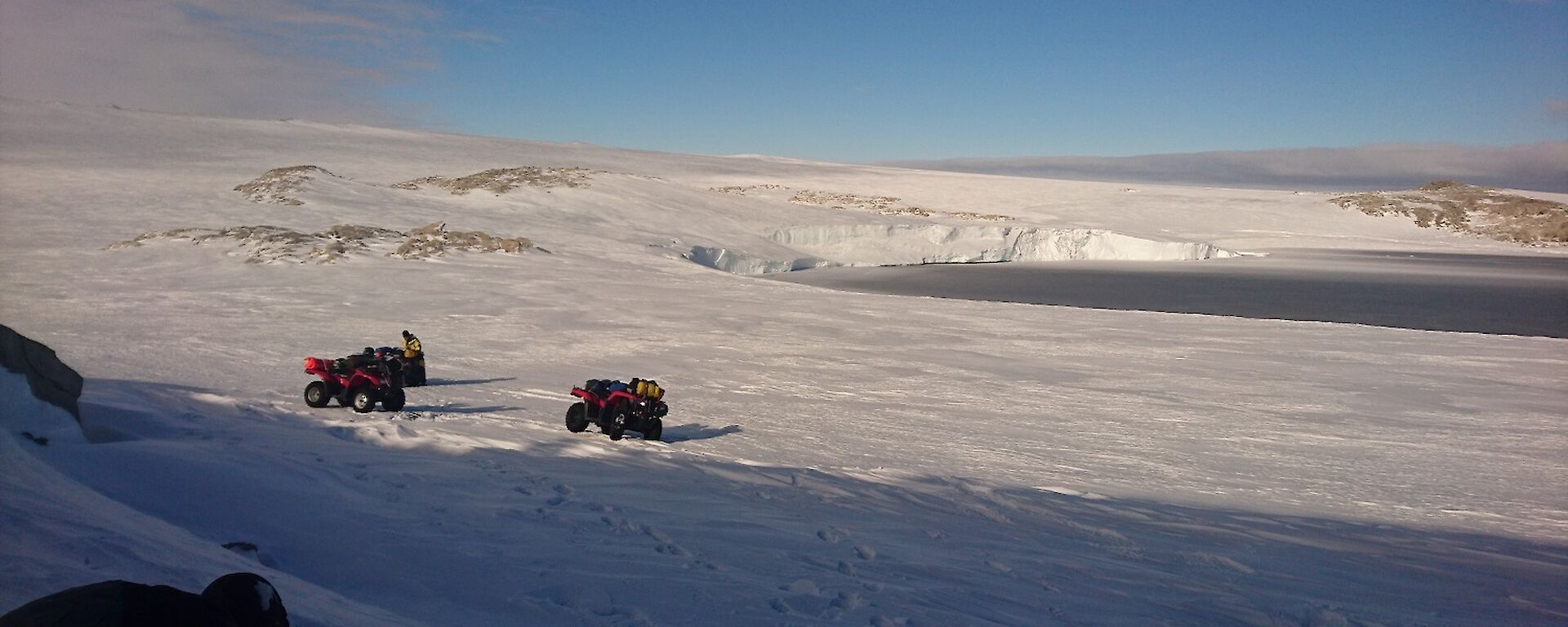 Snowy slope down to bay of blue water with three quad bikes parked in middle distance