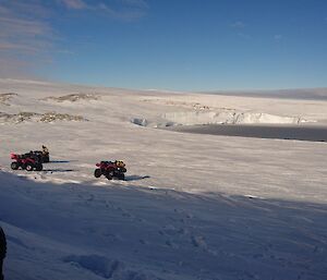 Snowy slope down to bay of blue water with three quad bikes parked in middle distance