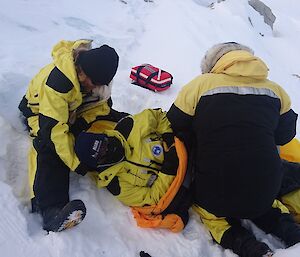 Man lying in snow with one man supporting head and another at hips rolling onto mat