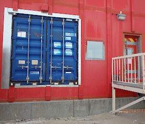 Side of the red building with side of blue shipping container inserted into the building as an opening and a small door with porch.