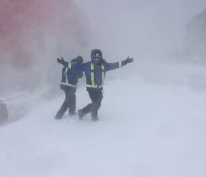 Two men in winter gear in centre picture with gale blowing around them with red building barely visible in the background