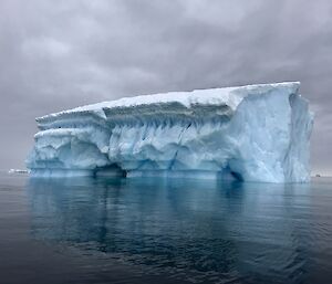 Large tabular iceberg with blue tinged side, reflection in grey calm water and behind overcast grey sky