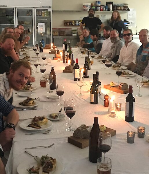 Large long table set with white tableclothes and candles, with expeditioners sitting down either side smiling towards camera.
