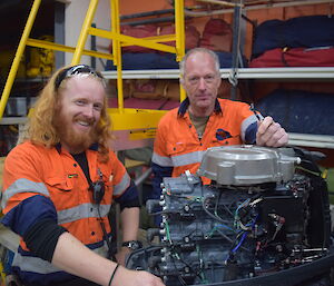 Two men in high vis shirts working on outboard motor and smiling towards camera