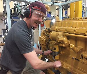 Expeditioner in t-shirt, beanie, ear muffs and safety glasses smiles at camera as working with spanner on main powerhouse engine