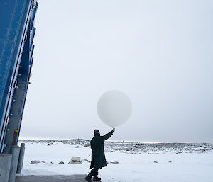 Man stepping out of blue balloon shed wearing overcoat and goggles and carrying large white balloon