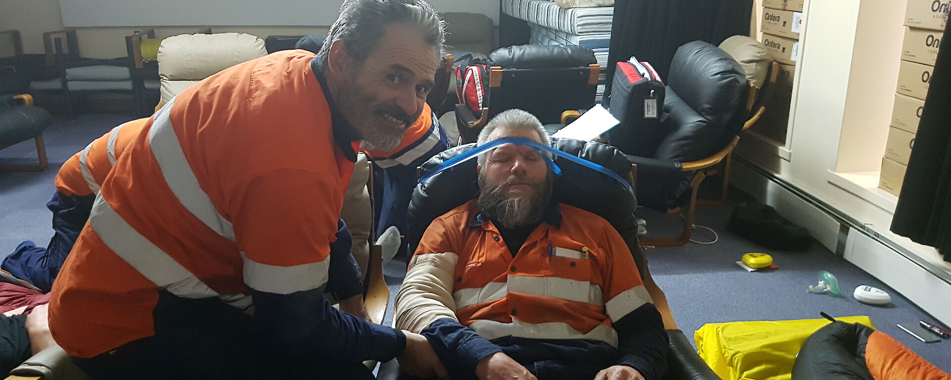 Patient unconscious in chair has blue tape around forehead and chair to prevent any movement, 1st aid responder looks to camera smiling