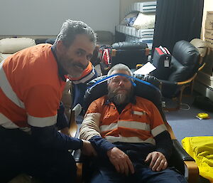 Patient unconscious in chair has blue tape around forehead and chair to prevent any movement, 1st aid responder looks to camera smiling