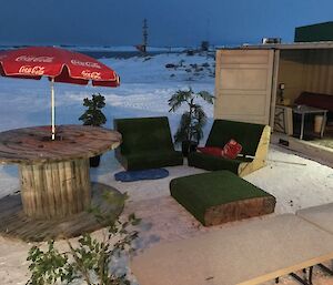 Shipping container with bbqs set up inside to right of picture, in centre picture outdoor chairs covered in fake turf, and large barrel with red sun umbrella installed. All on snow covered ground with Newcomb bay grey in the distance